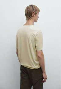 T-shirt in cotone beige con maniche corte e scollatura rotonda, mostrato dal retro. Indossato con pantaloni verde scuro. Texture liscia.