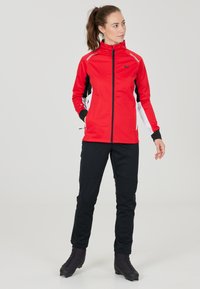 Giacca sportiva rossa con accenti neri e bianchi, colletto alto e tasche con zip, abbinata a pantaloni neri e calzature scure.