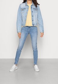 Žena v světlemodré denimové bundě, žlutém svetr, světlemodrých džínách a bílých teniskách, stojící proti jednoduché světle stěně.