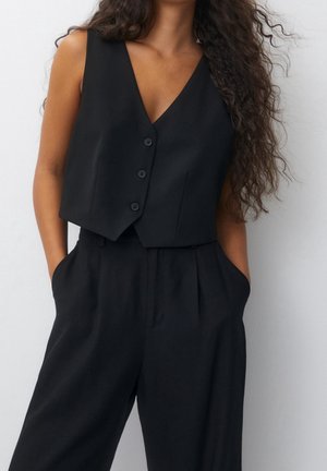 Femme portant un gilet noir sans manches boutonné et un pantalon noir taille haute, les mains dans les poches, debout devant un fond blanc.