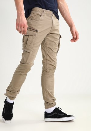 Homme portant un pantalon cargo beige avec poches zippées et à rabat, des baskets noires, des chaussettes blanches et une chemise bleu foncé, debout devant un fond uni.