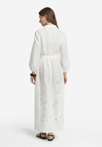 Lång vit linnekjol med bälte i midjan, har broderade blommönster i ryggen och långa puffärmar. Svarta sandaler synliga.