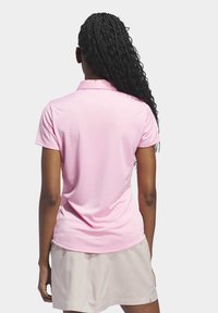 Femme aux longs cheveux tressés portant un polo à manches courtes rose clair et une jupe beige, vue de dos sur un fond uni.