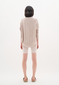 Svijetlosmeđi oversized pulover s dugim rukavima, uparen sa svijetlim šortsem. Model nosi cipele na slip. Pozadina je obična bijela.