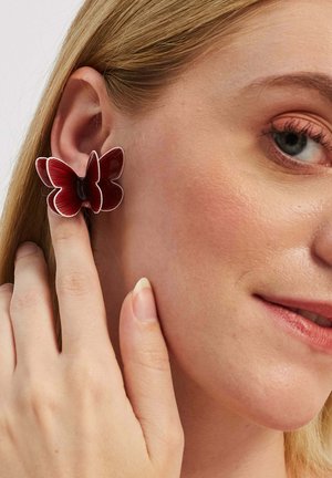 Sarkani tauri līdzīgi auskari, izgatavoti no glancēta materiāla ar smalku ribotu tekstūru un melnu centru, tos valkā modele, kura pieskaras ausij.