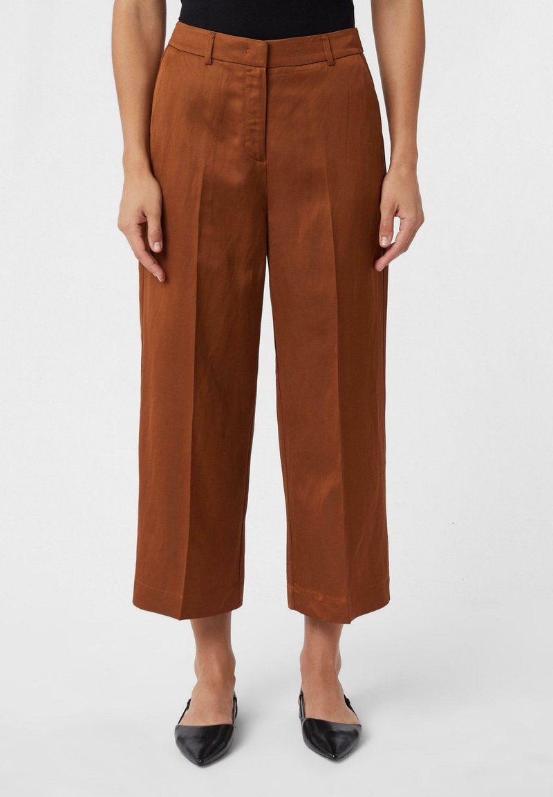 Pantalons larges taille haute marron en tissu doux, avec des plis avant et un ourlet propre. Associés à des chaussures noires à bout pointu.