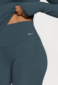 Legginsy Nike z wysokim stanem w ciemnym turkusie, wykonane z elastycznego materiału. Posiadają szare logo na pasku oraz płaskie szwy dla większego komfortu.