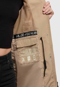 Interior de chaqueta beige con forro de malla, bolsillos etiquetados para pasaporte, teléfono y accesorios, con detalles de cordón y elástico de marca.