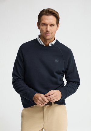 Uomo che indossa una felpa blu navy sopra una camicia a righe con colletto e pantaloni beige, in piedi contro uno sfondo chiaro uniforme, aggiustando la cintura.