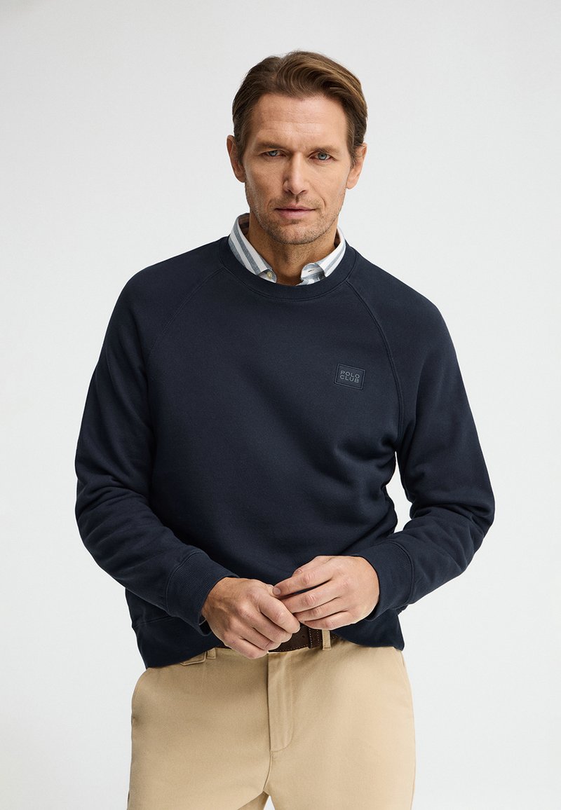 Hombre con sudadera azul marino sobre camisa de rayas con cuello y pantalones beige, de pie frente a un fondo claro y liso, ajustándose el cinturón.