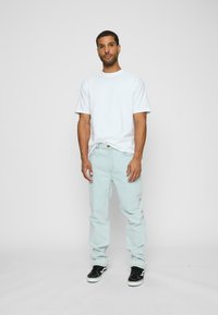 Jeans din bumbac de culoare albastru deschis, tricou alb cu mânecă scurtă și pantofi de skate negri. Modelul stă în picioare pe un fundal alb.