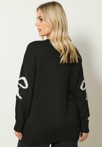 Czarna dziergana sweter o luźnym kroju, z długimi rękawami i kontrastowym białym napisem na rękawach. Teksturowana, miękka tkanina z ściągaczami.