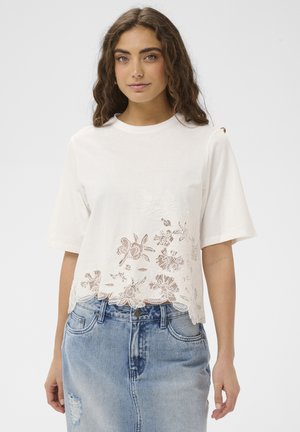 Mlada ženska z dolgimi kodrastimi lasmi, oblečena v belo kratkoročno majico s cvetličnimi čipkami in svetlo modro denim krilo.