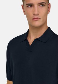 Jeune homme portant un polo à manches courtes bleu marine foncé, regardant légèrement sur le côté sur un fond clair uni.