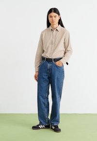 Beige långärmad skjorta med knappar, lösa blå denimjeans och svarta sneakers med vita ränder, står på grön golv mot en vit vägg.