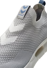 Jasnoszary dzianinowy sneakers wsuwany z białymi akcentami, teksturowaną cholewką oraz niebieską metką z wzorem w jodełkę.