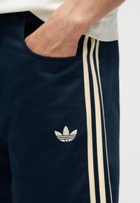 Χέρι στην τσέπη μπλε σκούρων παντελονιών Adidas με μπεζ ρίγες και λογότυπο Adidas trefoil στο μηρό.