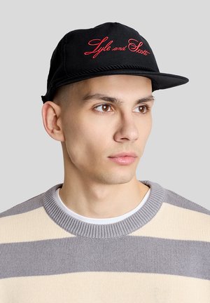 Ung mand med brune øjne iført en sort kasket med broderet "Lyle and Scott" og en grå- og beigefarvet stribet sweater, der kigger mod højre.