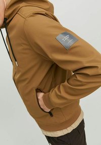 Hoodie zippé marron en tissu doux, avec une poche avant, un capuchon réglable et une étiquette rectangulaire sur la manche gauche avec des coordonnées.