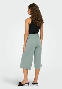 Jasnozielone kuloty z elastycznym paskiem, w długości do kostek, o gładkiej fakturze, zestawione z czarnymi klapkami na obcasie z kwiatowym akcentem.