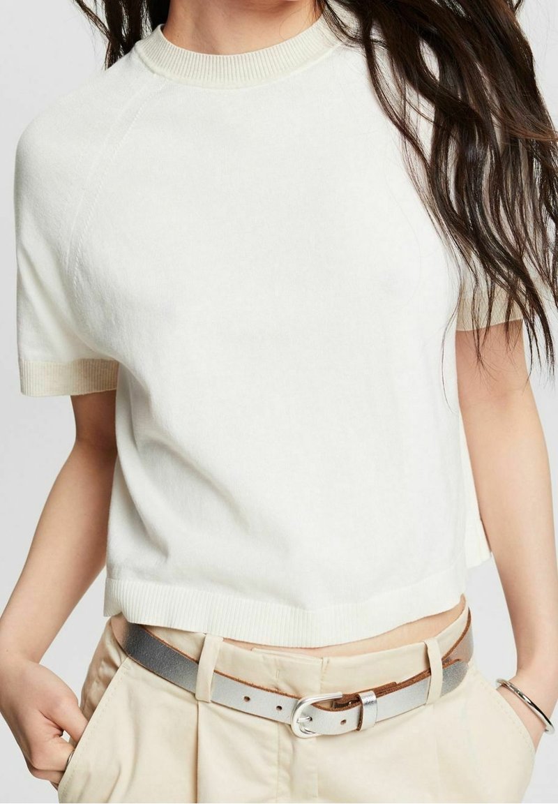 Kobieta w kremowej, krótkim rękawku dzianinowej bluzce włożonej w beżowe spodnie z wysokim stanem, z srebrnym paskiem, z rękami w kieszeniach i długimi, ciemnymi włosami.