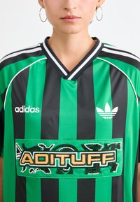 Žalios ir juodos vertikaliai dryžuotos marškinėlių su V formos iškirpte, su dideliu išsiuvinėtu "ADITUFF" užrašu ir baltu Adidas logotipu ant krūtinės.