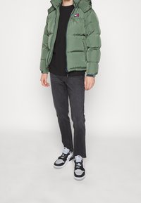 Chaqueta acolchada verde con capucha, detalles en azul marino, cierre de cremallera y parche con logo. Combinada con jeans negros y zapatillas negras/blancas.