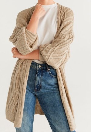 Personne portant un cardigan beige à torsades sur une chemise blanche et un jean bleu, les bras croisés et la main près du menton.