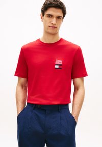 Άνδρας που φοράει κόκκινο T-shirt Tommy Hilfiger με λογότυπο στο στήθος και μπλε παντελόνι υφής, χέρια στις τσέπες, σε λευκό φόντο.