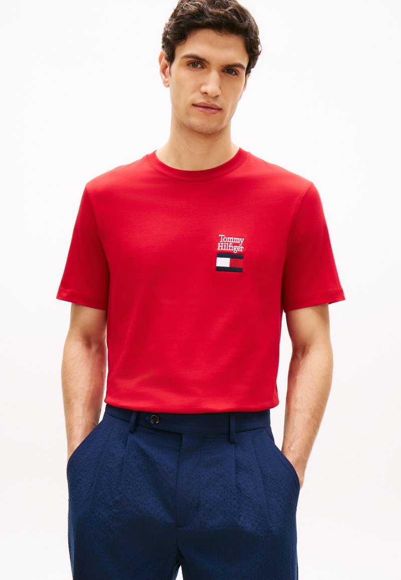 Άνδρας που φοράει κόκκινο T-shirt Tommy Hilfiger με λογότυπο στο στήθος και μπλε παντελόνι υφής, χέρια στις τσέπες, σε λευκό φόντο.