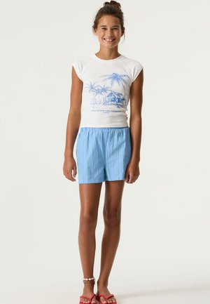 Chemise blanche à manches courtes avec graphique d'arbre palmier, associée à un short bleu avec des rayures verticales vertes. Sandales rouges vives. Coupe décontractée.