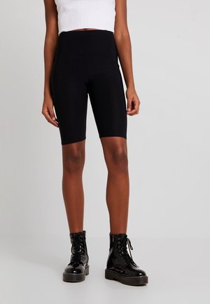 Persona con shorts negros de ciclista de talle alto, top blanco corto de punto acanalado y botas de combate negras brillantes con cordones, de pie contra un fondo liso.