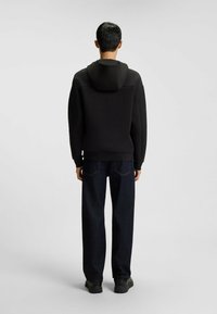 Felpa nera con cappuccio, dal taglio comodo, in tessuto testurizzato e con polsini a coste. Abbinata a jeans dritti blu scuro e scarpe nere.