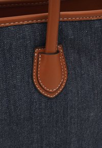 Jeanstotepåse med bruna läderdetaljer och sömmar. Har ett slätt handtag och en strukturerad design med en mörkblå textur.