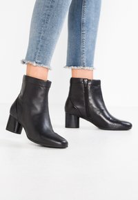 Stivaletti alla caviglia in pelle nera con punta appuntita, cerniera laterale e tacco largo. Il design presenta una superficie liscia e una parte superiore aderente.