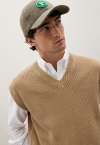 Maglione senza maniche beige lavorato a maglia con scollo a V, indossato sopra una camicia bianca con colletto, accompagnato da un cappellino a spina di pesce con una toppa verde.