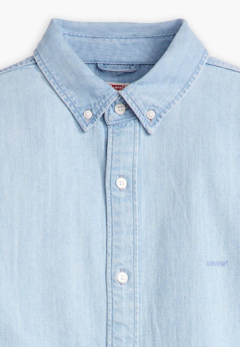 Chemise en denim bleu clair avec un col boutonné, présentant des boutons blancs et un subtil logo brodé sur la poitrine. Texture lisse.
