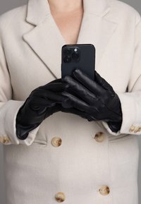 Schwarze Lederhandschuhe, die von einer Person getragen werden, die ein schwarzes Smartphone hält, kombiniert mit einem cremefarbenen Mantel mit großen Knöpfen.