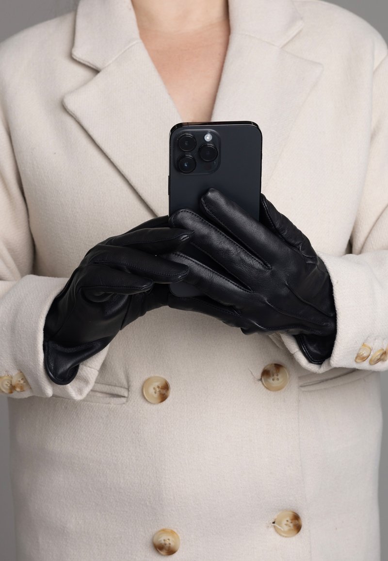 Schwarze Lederhandschuhe, die von einer Person getragen werden, die ein schwarzes Smartphone hält, kombiniert mit einem cremefarbenen Mantel mit großen Knöpfen.