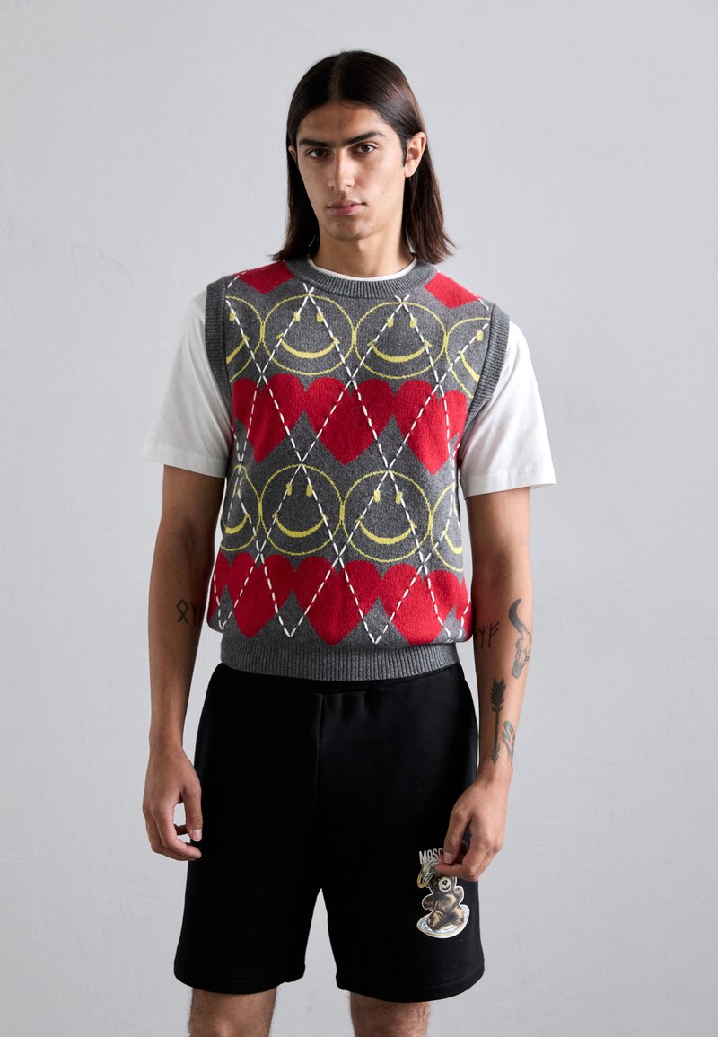 Junger Mann mit langen Haaren trägt einen grauen ärmellosen Pullover mit roten Herzen und gelben Smileys, ein weißes T-Shirt und schwarze Shorts mit einem Bärenpatch.