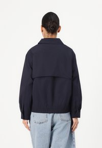 Nachtblauwe jas met een hoge kraag, elastische tailleband en pofmouwen. Heeft een gelaagd ontwerp aan de achterkant en een gladde, matte textuur.