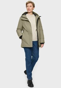 Vrouw in een beige winterjas met capuchon, een crèmekleurige gebreide trui, blauwe spijkerbroek en zwarte laarzen, staand met een hand in de zak.