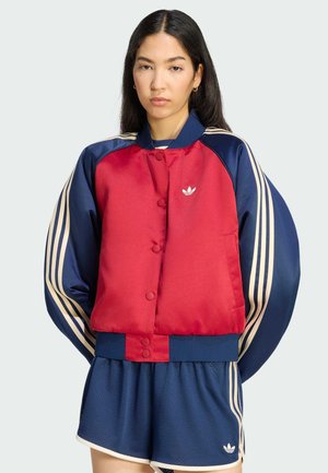 Giovane donna con lunghi capelli scuri che indossa una giacca sportiva rossa e blu navy e pantaloncini blu navy con strisce beige su uno sfondo chiaro.