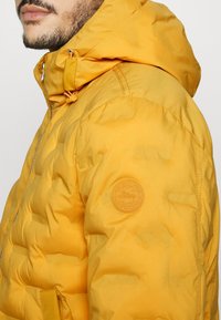Gul puffjacka med huva, med kviltat mönster och en rund logopatch på vänster ärm. Dragkedja och snörningsdetaljer synliga.