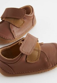 Chaussures en cuir marron avec une pièce en tissu texturé beige, fermeture à bande Velcro, bout arrondi et semelle en caoutchouc flexible.