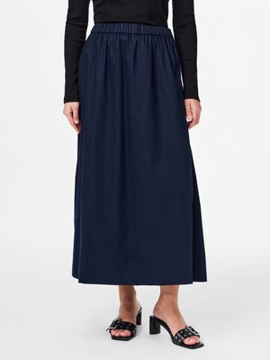 Kvinde iført en mørkeblå nederdel til anklerne med elastisk talje, sort langærmet top og sorte hælede sandaler med spænder.