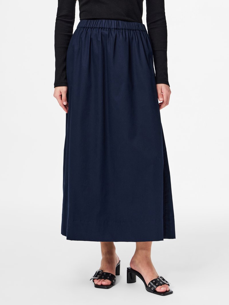 Γυναίκα που φοράει μπλε σκούρο, μακρύ μέχρι τον αστράγαλο φούστα με ελαστική μέση, μαύρο μπλουζάκι με μακριά μανίκια και μαύρα σανδάλια με τακούνια και αγκράφες.