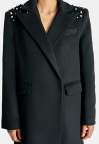 Cappotto di lana nera con spalle strutturate, decorato con borchie e cristalli, dotato di un colletto a scialle e due tasche frontali.