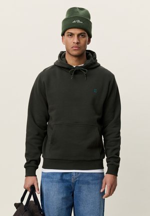 Jeune homme portant un bonnet vert et un sweat à capuche sombre, tenant un sac noir, debout devant un fond clair uni.