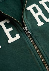Gros plan d'une veste verte zippée avec des lettres blanches brodées et un curseur de fermeture éclair en métal portant la mention « DRY ».